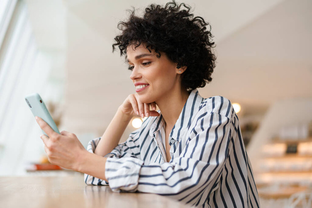 Beautiful cheerful curly woman using mobile phone while sitting in cafe - Photo, Image