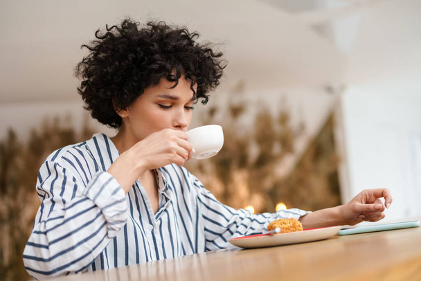 Beautiful pleased curly woman eating cake while drinking coffee in cafe - Photo, image