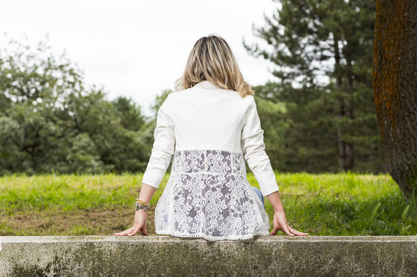 Blond włosy kobieta siedzi plecami do kamery na kamiennej ławce w parku w Asturias, Hiszpania.Kobieta ma na sobie dżinsy i białą kurtkę. Zdjęcie produktu mody zrobione w pochmurny dzień na zewnątrz. - Zdjęcie, obraz