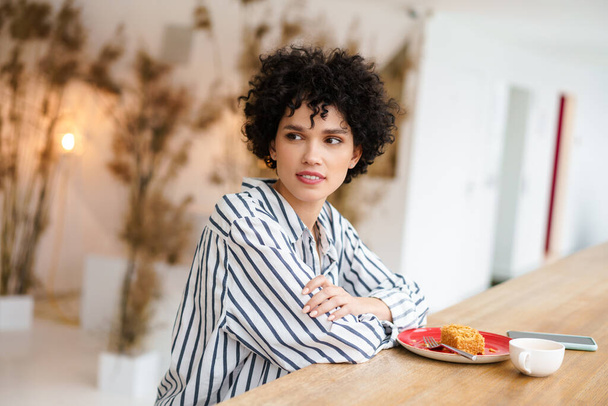 Beautiful caucasian curly woman drinking coffee with cake while sitting in cafe - 写真・画像