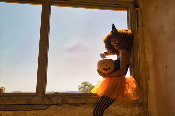Mujer latina joven vestida como una bruja y sentada en un marco de ventana riendo en voz alta y metiéndose en una calabaza de Halloween. - Foto, Imagen