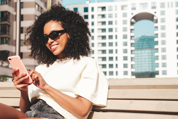 Beautiful black woman with afro curls hairstyle.Smiling model in white hoodie. Sexy carefree female posing on the street background in sunglasses. Looking at smartphone screen, using apps - Φωτογραφία, εικόνα
