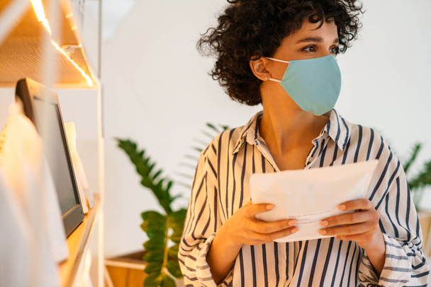 Curly focused woman in face mask examining product in shop - Photo, image
