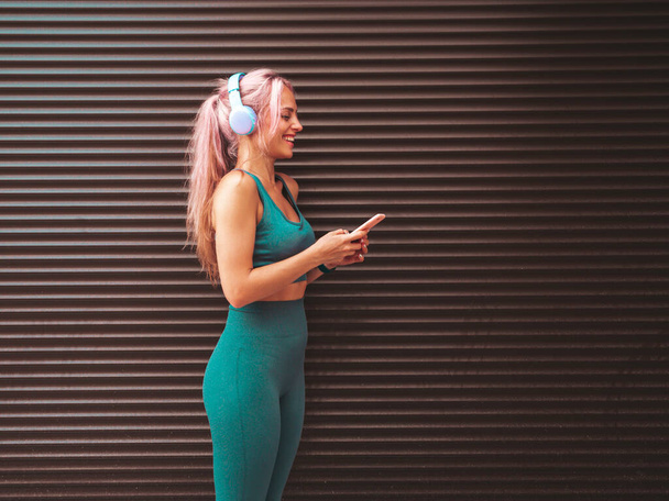 Fitness smiling woman in green sports clothing with pink hair. Young beautiful model with perfect body.Female in the street near roller shutter wall.Listening music in wireless headphones - Φωτογραφία, εικόνα