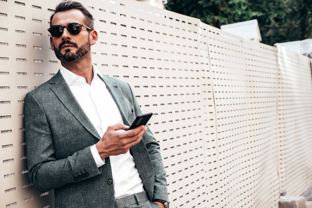 Portrait of handsome confident stylish hipster lambersexual model.Modern man dressed in elegant suit. Fashion male posing in the street near fence at sunset. In sunglasses.Using smartphone apps. - Foto, imagen
