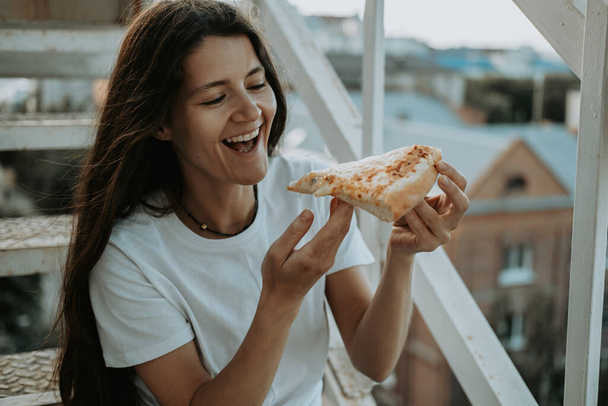 Young beautiful brunette is sitting on the stairs and eating pizza. Eating outdoor. - Fotó, kép