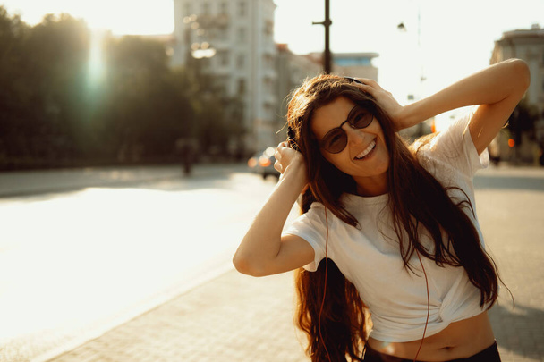 Beautiful happy young woman using cell phone and headphones and listening music in the city streets. Positive emotions, lifestyle expressive portrait in summer. - Zdjęcie, obraz