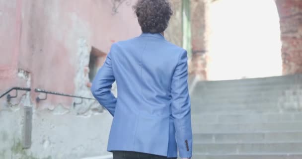 kıvırcık saçlı, mavi ceketli, bavullu ve Romanya 'nın ortaçağ eski şehrinde merdivenleri çıkan sıradan bir adam. - Video, Çekim