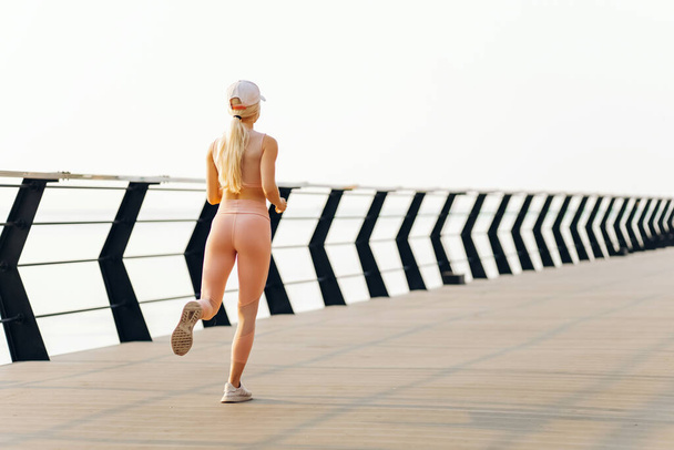 Young fitness woman running outdoors on the bridge, Sportswoman in sportswear runs in the morning with bright sunlight on the sea coast, Sports, motivation, healthy lifestyle - Fotografie, Obrázek