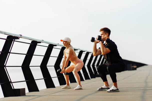 Fit young couple doing kettlebell squats, sporty muscular man and woman exercising outdoors on the coast at sunrise, Healthy lifestyle concept, sport - Foto, Bild