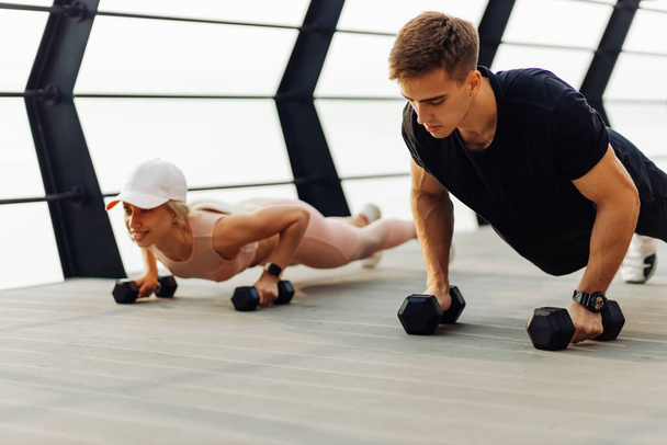 Young couple exercising outdoors, Attractive sportive woman and handsome muscular man are exercising on the bridge on the coast, Doing plank on kettlebell, doing push-ups, Sport concept, motivation - Φωτογραφία, εικόνα