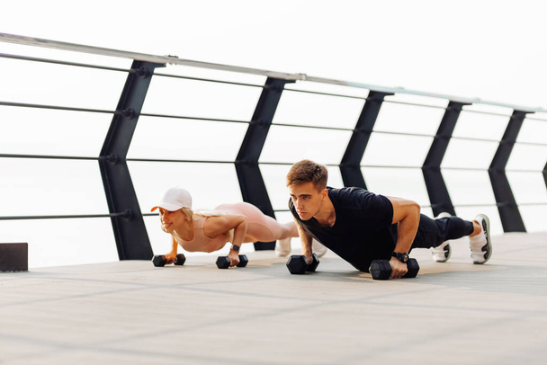 Young couple exercising outdoors, Attractive sportive woman and handsome muscular man are exercising on the bridge on the coast, Doing plank on kettlebell, doing push-ups, Sport concept, motivation - Foto, afbeelding