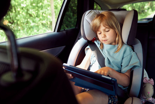 Little girl using digital tablet on rear seat of car                                - Фото, изображение
