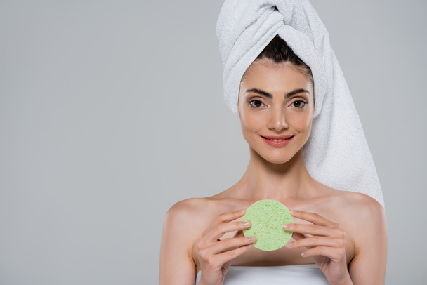 улыбающаяся молодая женщина с полотенцем на голове держа зеленую губку красоты изолированы на сером  - Фото, изображение