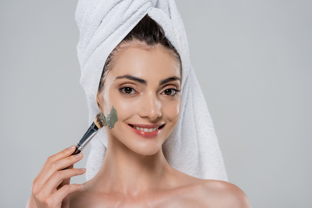 cheerful young woman with towel on head applying green clay mask with cosmetic brush isolated on grey - Photo, Image