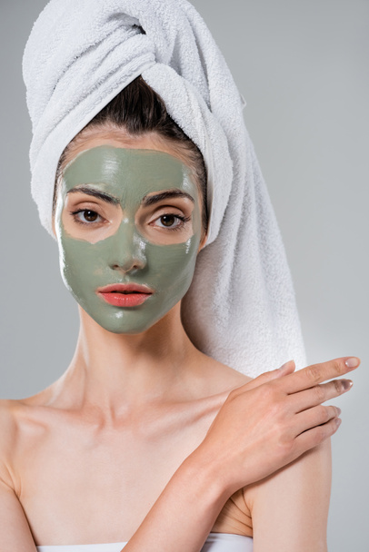young woman with towel on head and green clay mask on face touching bare shoulder isolated on grey - Foto, Imagem