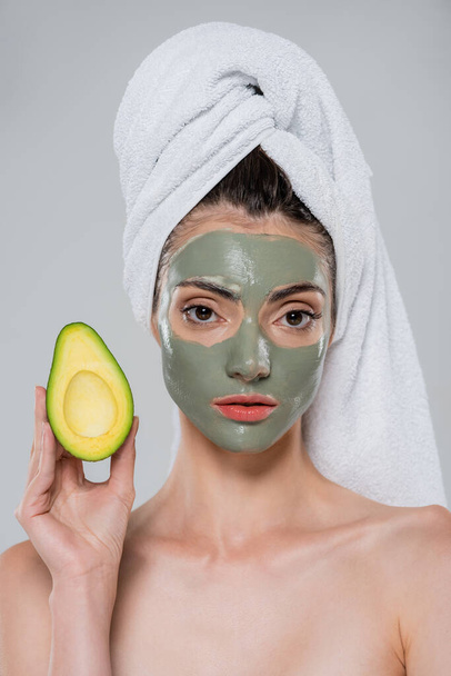 young woman with towel on head and green clay mask on face holding half of avocado isolated on grey - Photo, Image