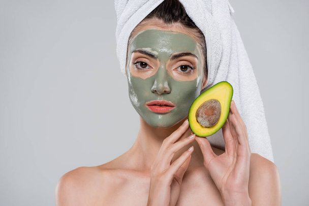 young woman with towel on head and green clay mask holding ripe avocado isolated on grey - Fotó, kép