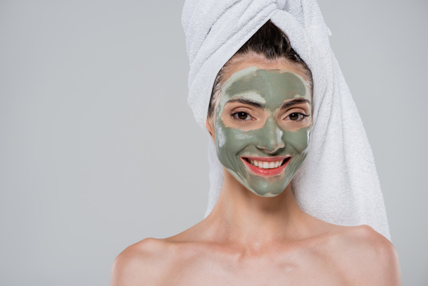 радостная молодая женщина с полотенцем на голове и зеленой глиняной маской на лице изолированы на сером - Фото, изображение