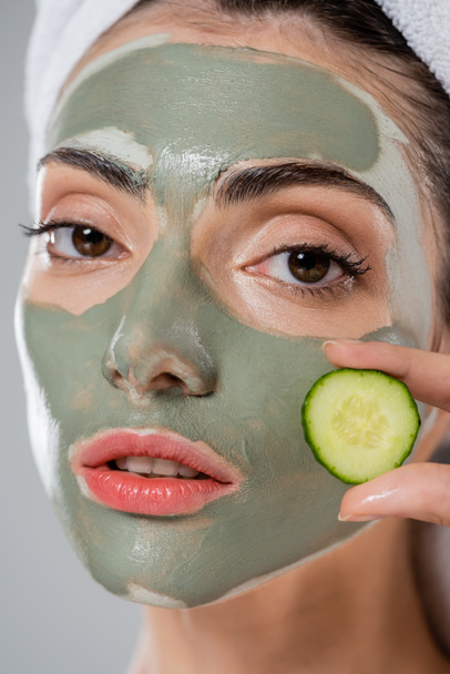 primo piano di giovane donna con maschera di argilla verde sul viso tenuta tagliata cetriolo isolato su grigio - Foto, immagini
