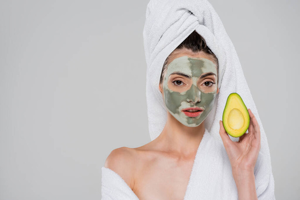 woman with towel on head and clay mask holding green avocado isolated on grey - Foto, Imagem