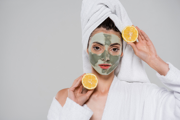 junge Frau mit grüner Lehmmaske, die reife Orangenhälften isoliert auf grau hält - Foto, Bild