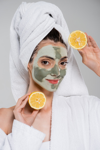 smiling young woman with green clay mask holding ripe orange halves isolated on grey - Foto, immagini