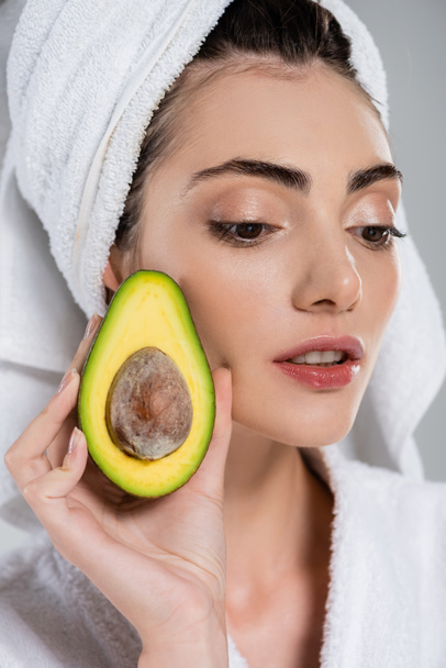 close up of young woman in bathrobe holding half of avocado isolated on grey - Fotoğraf, Görsel