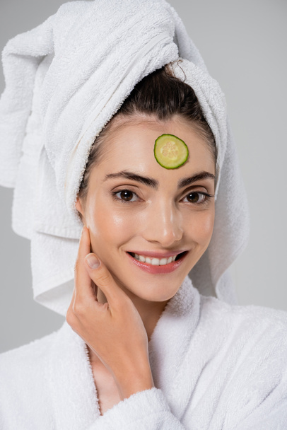 gelukkig jong vrouw met handdoek op hoofd en gesneden komkommer op gezicht geïsoleerd op grijs - Foto, afbeelding