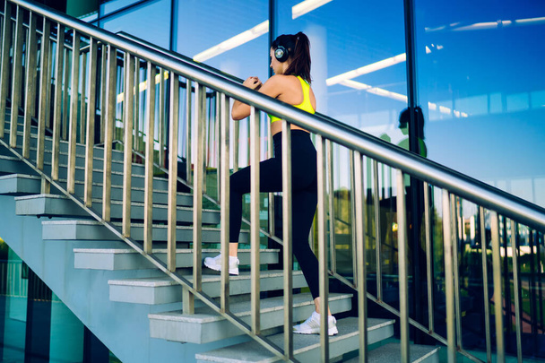 Young female in yellow top black leggings white sneakers and headphones doing exercises on stairway of modern urban building on sunny day - Фото, зображення