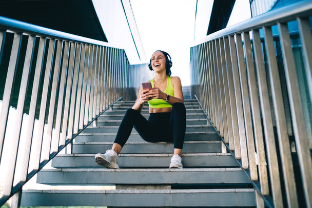 From below of cheerful slim female jogger in casual sportswear and sneakers sitting on concrete stairs of building and listening to music in headphones - Foto, imagen