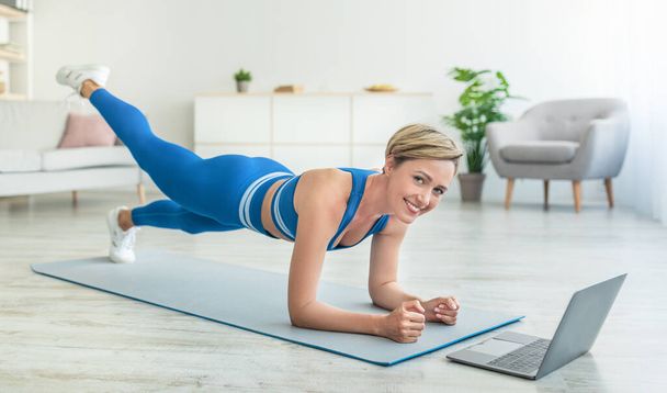 Femme heureuse faisant planche sur tapis avec ordinateur portable à la maison - Photo, image