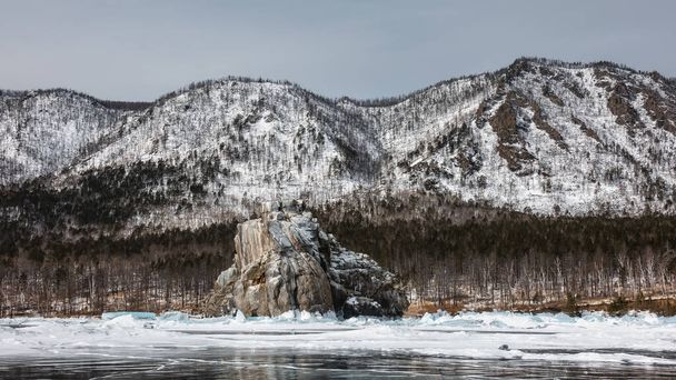 Une roche de granit aux contours bizarres et au sommet plat s'élève au-dessus d'un lac gelé. Il y a de la neige à la base, hummocks. Réflexion sur la glace. Le fond est une chaîne de montagnes enneigée. Baïkal - Photo, image