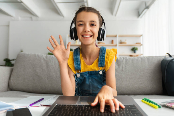 Girl in headphones using laptop, waving hand to webcam - Фото, изображение