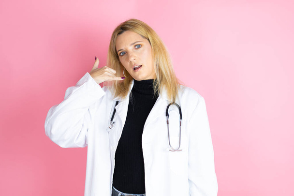 Joven doctora rubia usando estetoscopio de pie sobre un fondo rosa aislado confundida haciendo gesto telefónico con la mano y los dedos como hablar por teléfono - Foto, imagen