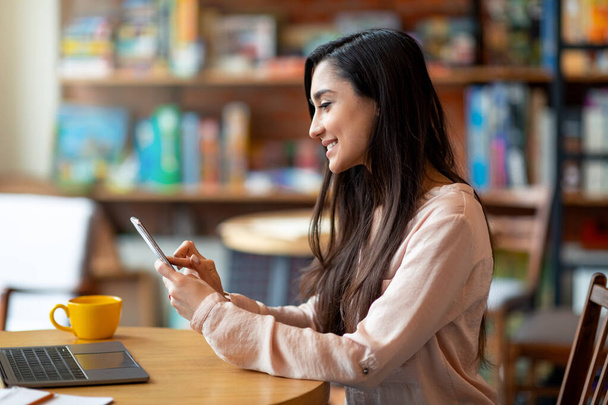 Heureuse femme latine tenant un téléphone portable et utilisant un ordinateur portable, pigiste travaillant au café, espace de copie, vue latérale - Photo, image