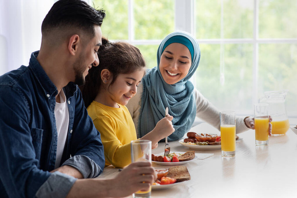 Обід разом. Ісламські батьки і маленька донька, які отримують задоволення від смачної їжі на кухні - Фото, зображення