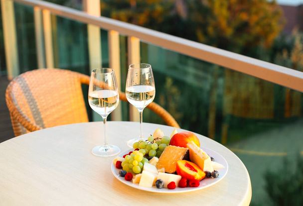 Tagli di formaggio assortiti e piatto di frutta da due bicchieri di vino bianco sul tavolo rotondo in piedi sul balcone dell'hotel con vista sulla foresta estiva al tramonto. Spuntini leggeri per coppia in vacanza - Foto, immagini