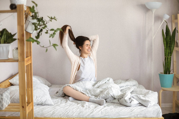 Νεαρή ευτυχισμένη χαλαρή γυναίκα στην πιτζάμα κάθεται στο κρεβάτι με κλειστά μάτια το πρωί στο σπίτι, τεντώνοντας το σώμα της και χαμογελώντας μετά από να ξυπνήσει, απολαμβάνοντας νέα όμορφη μέρα. Έννοια αφύπνισης - Φωτογραφία, εικόνα