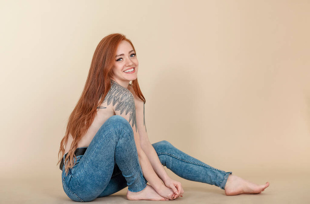 Młoda uśmiechnięta piękna kobieta z długimi rudymi włosami patrzy w kamerę z uśmiechem na twarzy, siedzi bokiem w dżinsach półnaga w studio, pokazując ramię z tatuażem skrzydła - Zdjęcie, obraz