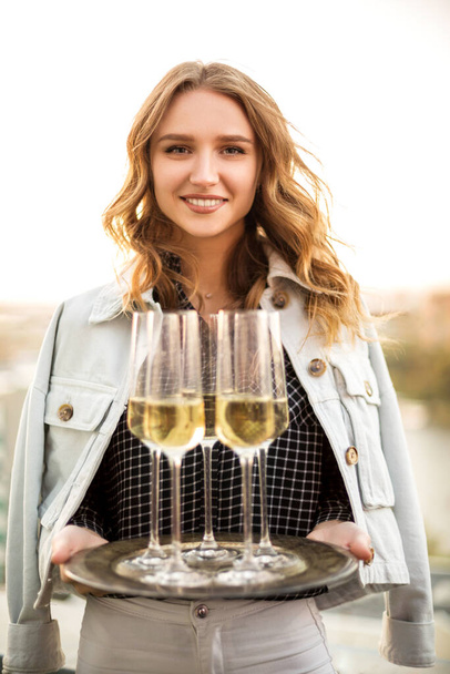 Young blonde smiling woman holding tray with champagne sparkling wine in flute glasses, standing outdoors over blurred countryside background, celebrating birthday party on cool summer evening - 写真・画像
