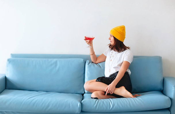 Обрезанный снимок молодой улыбающейся женщины с бокалом коктейля в руке и красным спиртным напитком, сидящим дома на диване, одетой в модную одежду и аксессуары, наслаждающейся вечеринкой с друзьями в помещении - Фото, изображение