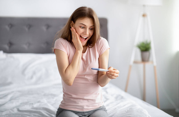 Jeune femme excitée vérifiant son récent test de grossesse, se sentant heureuse sur la maternité future sur le lit à la maison - Photo, image