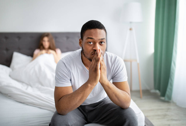 Молодой черный парень чувствует себя несчастным, сидя на кровати глубоко в мыслях, его разочарованная жена на заднем плане - Фото, изображение