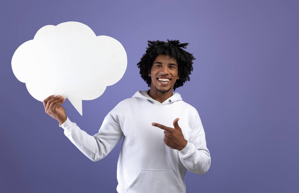 Funky black teen guy pointing at empty speech bubble, smiling at camera on violet studio background, mockup - Photo, Image