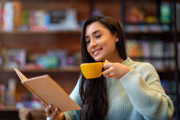 Nuori arabinainen lukemassa suosikkikirjaa ja nauttimassa kahvista kahvilassa, istumassa kahvilassa ja viettämässä aikaa nauttien - Valokuva, kuva