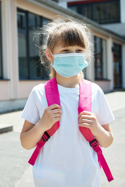 Retrato de uma menina com uma mochila em uma máscara médica no pátio da escola. Proteção de crianças e crianças em idade escolar durante o período de vírus e epidemias de gripe. Foto vertical - Foto, Imagem