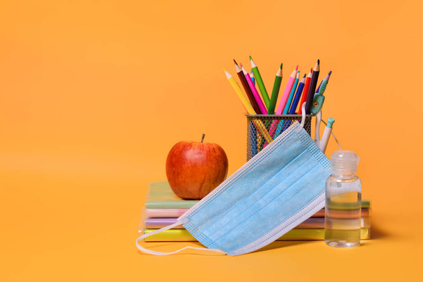 Schulbedarf, ein Apfel, medizinische Masken, Desinfektionsmittel auf orangefarbenem Hintergrund. Zurück zur Schule während der Coronavirus COVID-19 Pandemie. Kopierraum. - Foto, Bild