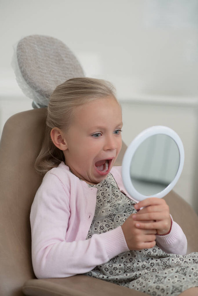Nettes blondes Mädchen schaut ihre Zähne im Spiegel an - Foto, Bild