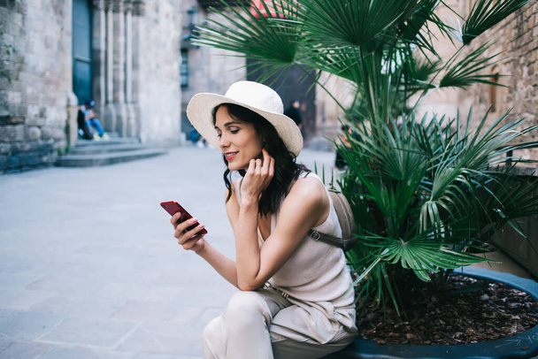 Femme souriante dans le chapeau et avec sac à dos assis et regardant l'écran du smartphone sur fond de bâtiments anciens en briques - Photo, image